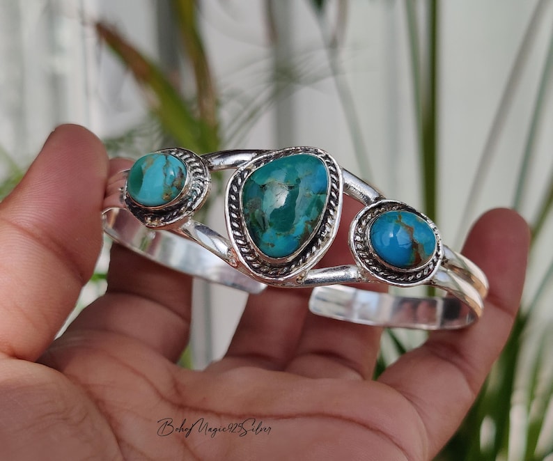 May include: A silver cuff bracelet featuring three turquoise stones. The stones are oval and teardrop-shaped, set in ornate silver bezels. The bracelet has a polished finish and a hinged design.