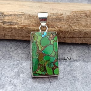 May include: A silver pendant with a rectangular green and brown stone. The stone has a mottled pattern and is set in a silver frame. The pendant is on a wooden surface.
