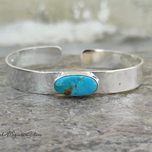May include: A silver cuff bracelet with a hammered texture. The bracelet features an oval turquoise stone in the centre. The turquoise stone has brown and blue hues. The bracelet is open at the ends.