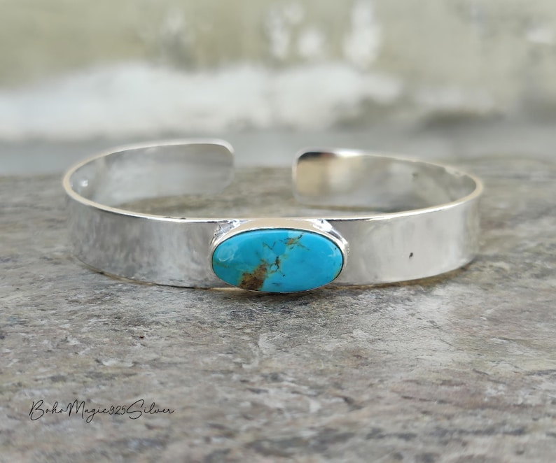 May include: A silver cuff bracelet with a hammered texture. The bracelet features an oval turquoise stone in the centre. The turquoise stone has brown and blue hues. The bracelet is open at the ends.