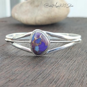 May include: A sterling silver cuff bracelet featuring a central teardrop-shaped purple and blue stone with copper veining. The band has a split design with a twisted rope texture detail surrounding the stone. The text Boho Magic 925 Silver is visible.