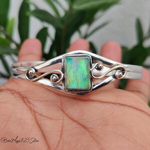 May include: A silver cuff bracelet featuring a rectangular, iridescent green opal stone set in the center. The metal band displays decorative swirling wire details on both sides of the stone. The text "Boho Magic 925 Silver" is printed in the corner.