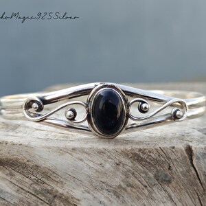 May include: A silver bracelet with a black oval gemstone set in a delicate, swirling design. The bracelet is made of 925 sterling silver.