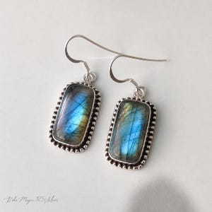 Puede incluir: Un par de pendientes de plata con piedras rectangulares de labradorita. Las piedras muestran tonos iridiscentes azules y verdes. Cada pendiente tiene un borde con cuentas y un cierre de gancho. Los pendientes se muestran sobre una superficie blanca.