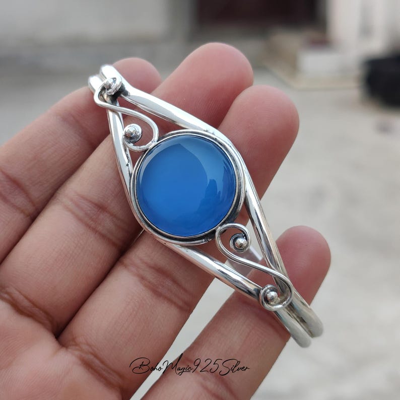 May include: A silver bangle bracelet with a blue gemstone set in a decorative silver frame. The bracelet has a delicate, intricate design.