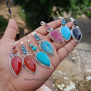 May include: A collection of gemstone pendants in various colors, including red, turquoise, and black. Each pendant has a teardrop shape and is set in a silver frame. The pendants are displayed on a hand.