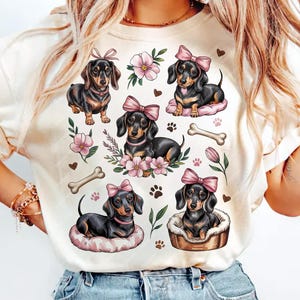 Op de afbeelding: Crèmekleurig T-shirt met een herhaald patroon van teckels met roze strikken, bloemen, pootafdrukken en hondenbotten. De teckels zijn zwart en bruin. Het shirt is gemaakt van een zacht materiaal.