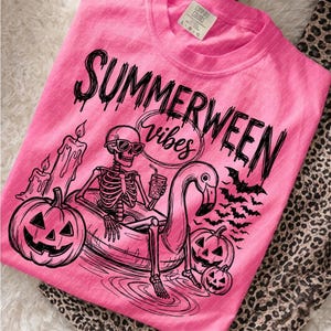 May include: A bright pink t-shirt featuring a black graphic print of a skeleton wearing sunglasses, sitting in a flamingo pool float. The design includes pumpkins, candles, bats, and the text "SUMMERWEEN vibes" in a stylized, dripping font.