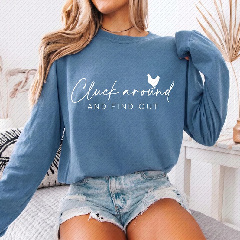 May include: A long-sleeved, blue sweatshirt with the phrase "Cluck around AND FIND OUT" in white script. A small white chicken graphic is to the right of the word "around". The sweatshirt is paired with denim shorts.