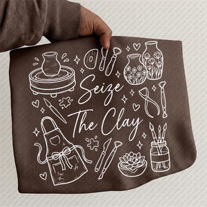 Puede incluir: Un bolso tote marrón con un dibujo lineal blanco de herramientas de cerámica y la frase "Seize The Clay". El diseño incluye un torno de alfarero, pinceles, un delantal y una suculenta. El bolso lo sostiene una persona.