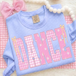 Op de afbeelding: Een lichtblauwe sweatshirt met het woord "DANCE" in roze ruitjesletters, versierd met witte madeliefjes en gele strikken. De sweatshirt is opgevouwen op een roze en wit geruit oppervlak, met een roze strik en een witte kralenketting op de achtergrond.