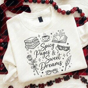 May include: Cream-colored sweatshirt with a black graphic design featuring books, a cup of coffee, a masquerade mask, and the text "Spicy Pages & Sweet Dreams." The design includes a rose, glasses, and a feather.