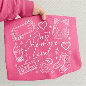 Puede incluir: Una manta rosa con ilustraciones blancas de temática gaming, que representa una consola portátil, auriculares, un mando y calcetines. El texto "Just One More Level" está escrito en una fuente de escritura. La manta está sujeta por una persona que lleva un jersey rosa.