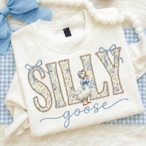 May include: White sweatshirt featuring the words "SILLY goose" in a playful font. The letters are adorned with a blue gingham design and a goose illustration. Blue bows decorate the letters. The sweatshirt rests on a blue and white checked background.