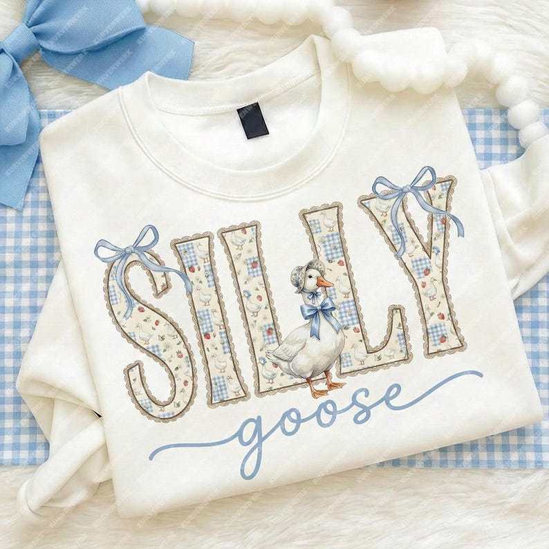 May include: White sweatshirt featuring the words "SILLY goose" in a playful font. The letters are adorned with a blue gingham design and a goose illustration. Blue bows decorate the letters. The sweatshirt rests on a blue and white checked background.