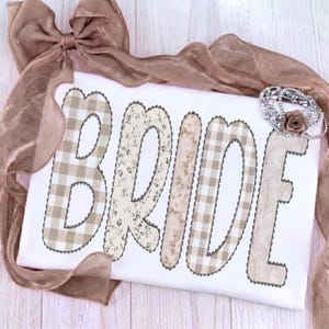May include: A white decorative pillow with the word "BRIDE" in large patchwork letters. The letters feature floral, plaid, and solid patterns in neutral tones, with a scalloped border. A brown ribbon and a pearl bracelet with a rose accent the pillow.
