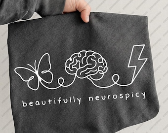 Könnte beinhalten: Ein dunkelgraues T-Shirt mit einer durchgehenden weißen Strichzeichnung eines Schmetterlings, eines menschlichen Gehirns und eines Blitzes. Unter der Grafik steht der Text "beautifully neurospicy" in einer einfachen weißen Schrift.
