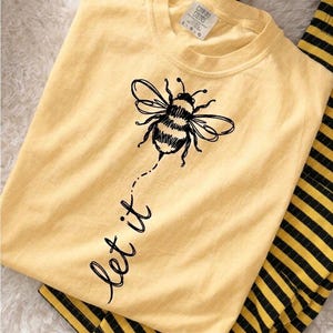 May include: A pale yellow t-shirt featuring a black line-art illustration of a bumblebee centered on the chest. Below the bee, the text "let it" is printed in a black cursive script. The shirt has a crew neckline and a Comfort Colors brand label.