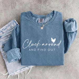 May include: A blue-grey sweatshirt with the phrase "Cluck around AND FIND OUT" in white script, along with a white chicken graphic. The sweatshirt is folded with the sleeves cuffed. A silver necklace with a round pendant is on top.