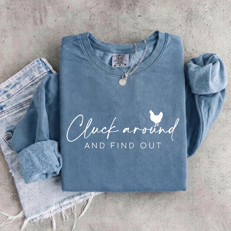 May include: A blue-grey sweatshirt with the phrase "Cluck around AND FIND OUT" in white script, along with a white chicken graphic. The sweatshirt is folded with the sleeves cuffed. A silver necklace with a round pendant is on top.
