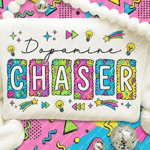 Può includere: Una felpa bianca con la scritta "Dopamine CHASER" in un font colorato e fumettistico. Il design include fulmini, stelle, lampadine e faccine sorridenti, su uno sfondo rosa e giallo vivace.