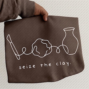 May include: A brown apron with a white line drawing of pottery tools and a vase. The text "seize the clay" is printed in white at the bottom. The apron is held by a person.