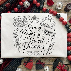 May include: White t-shirt with a black graphic design featuring books, a cup of coffee, a rose, and the text "Spicy Pages & Sweet Dreams." The design includes a burning book, a masquerade mask, glasses, and a feather.