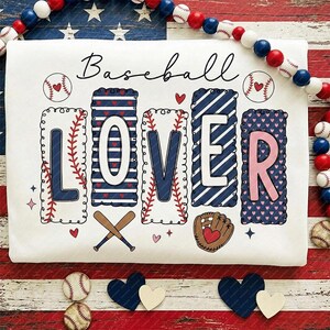 May include: White t-shirt with the words "Baseball Lover" in a decorative design. The letters are in blue and pink, with baseball-themed graphics. The shirt is decorated with baseballs, hearts, and baseball bats.