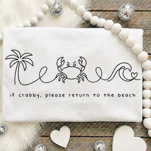 May include: White beach towel with a black line drawing of a palm tree, crab, wave, and heart. The text "if crabby, please return to the beach" is printed below the drawing. Decorative elements surround the towel.