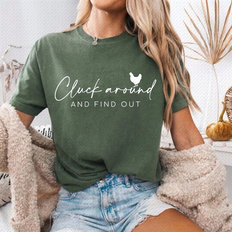 May include: Green t-shirt with the text "Cluck around AND FIND OUT" in white script and a small white chicken graphic. The shirt is a solid olive green color and has short sleeves.