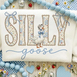 May include: White sweatshirt featuring the words "SILLY goose" in blue script. The letters are decorated with a floral and gingham pattern, and a cartoon goose wearing a bonnet is in the center. Blue bows accent the letters.