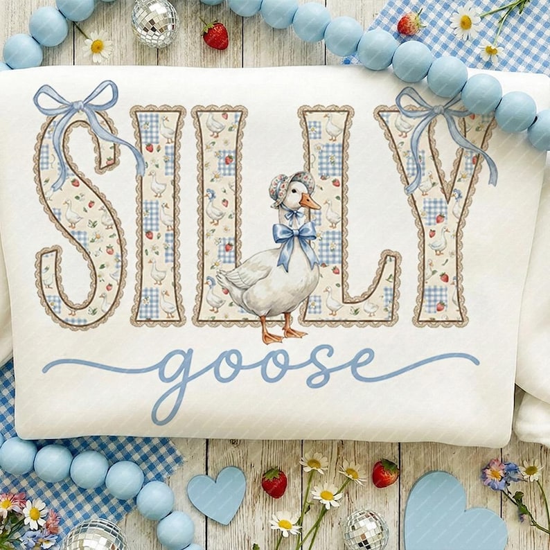 May include: White sweatshirt featuring the words "SILLY goose" in blue script. The letters are decorated with a floral and gingham pattern, and a cartoon goose wearing a bonnet is in the center. Blue bows accent the letters.