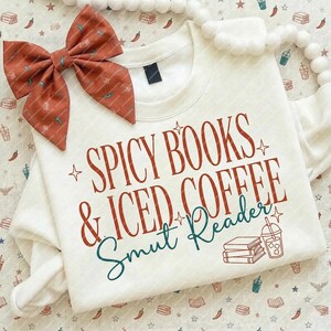 May include: Cream-colored sweatshirt with the text "Spicy Books & Iced Coffee Smut Reader" in red and teal. A matching bow with a chili pepper pattern is on the left. The design includes book and iced coffee graphics.
