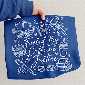 May include: A blue towel with white line art illustrations of a gavel, scales of justice, books, coffee cup, pen, and glasses. The text "Fueled By Caffeine & Justice" is written in a cursive font.