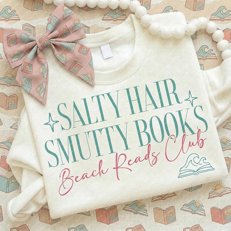 May include: Cream-colored sweatshirt with the text "SALTY HAIR SMUTTY BOOKS Beach Reads Club" in teal and pink. A matching bow with a book and sun pattern is on the left. A white beaded garland is on the right.