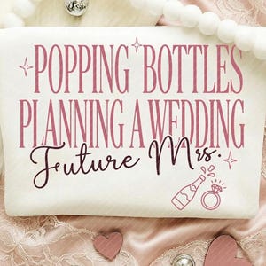 May include: A white cosmetic bag with the text "Popping Bottles Planning a Wedding Future Mrs." in pink lettering. A champagne bottle and diamond ring graphic are also featured. The bag is surrounded by decorative elements.