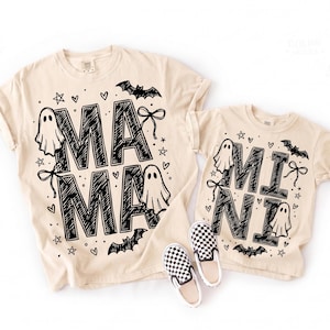 Puede incluir: Dos camisetas de color beige con estampados gráficos en negro. La camiseta más grande muestra el texto MAMA y la más pequeña el texto MINI, rodeados de ilustraciones de fantasmas, murciélagos, estrellas, corazones y lazos. Incluye un par de zapatos a cuadros.