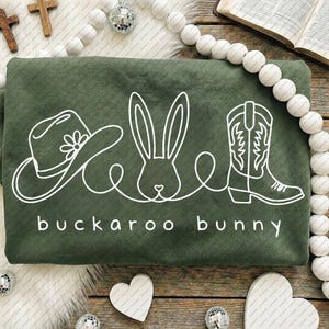 May include: An olive green sweatshirt with a white line drawing of a cowboy hat, a bunny, and a cowboy boot. The words "buckaroo bunny" are printed in white below the drawing. The sweatshirt is folded.