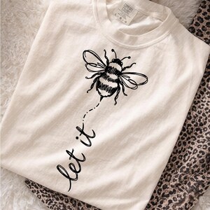 Op de afbeelding: Een cr&egrave;mekleurig T-shirt met korte mouwen en een zwarte lijntekening van een hommel. Onder de bij staat de tekst "let it" in cursief schrift gedrukt. Het shirt heeft een ronde hals en een Comfort Colors merklabel in de nek.