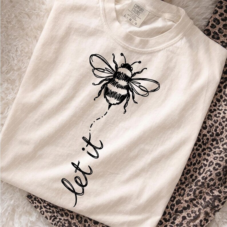 Op de afbeelding: Een cr&egrave;mekleurig T-shirt met korte mouwen en een zwarte lijntekening van een hommel. Onder de bij staat de tekst "let it" in cursief schrift gedrukt. Het shirt heeft een ronde hals en een Comfort Colors merklabel in de nek.