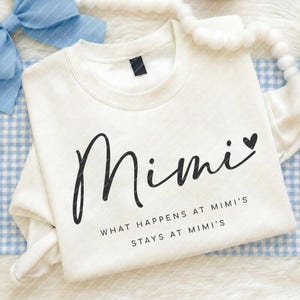 May include: Cream-coloured sweatshirt featuring the word "Mimi" in black script, with a heart symbol. Underneath, the text reads "WHAT HAPPENS AT MIMI'S STAYS AT MIMI'S" in a smaller, black font.