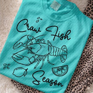 Può includere: Una t-shirt turchese con un'illustrazione a linee nere di un gambero di fiume, un limone e spicchi di limone. Il testo "Craw Fish Season" è stampato in corsivo nero. La maglietta presenta un'etichetta del marchio Comfort Colors sul collo.