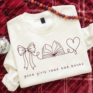 May include: Cream-colored sweatshirt with a dark brown line drawing of a bow, an open book, and a heart. The text "good girls read bad books" is printed below the graphic. A decorative beaded garland is draped across the top.