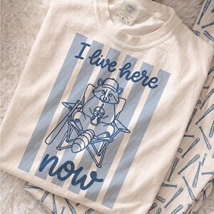 Può includere: T-shirt color crema con un design a righe blu e la scritta "I live here now". La grafica presenta un procione su una sedia a sdraio che tiene in mano una bevanda. Sono visibili pantaloni del pigiama abbinati con un motivo a mazza da baseball.