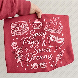 May include: A red decorative pillow with the text "Spicy Pages & Sweet Dreams" in white script. The pillow features white illustrations of books, a teacup, a rose, a masquerade mask, glasses, and a feather.
