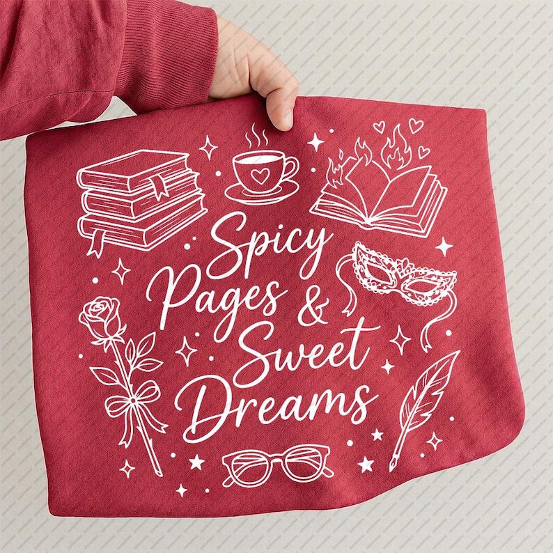 May include: A red decorative pillow with the text "Spicy Pages & Sweet Dreams" in white script. The pillow features white illustrations of books, a teacup, a rose, a masquerade mask, glasses, and a feather.