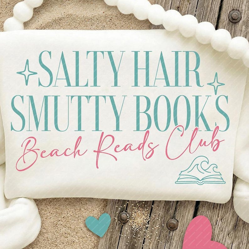 May include: A white beach towel with the text "SALTY HAIR SMUTTY BOOKS Beach Reads Club" in teal and pink. The towel is on a wooden surface with sand and decorative hearts.
