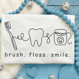 May include: White t-shirt featuring a black line drawing of a toothbrush, tooth, and dental floss. The words "brush. floss. smile." are printed below in a black, sans-serif font.
