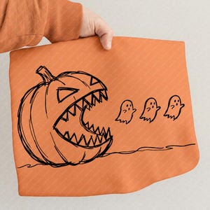 Pode incluir: Um artigo de tecido laranja com um desenho de linhas pretas de uma grande abóbora com uma boca larga e serrilhada aberta, como se fosse devorar três pequenas figuras de fantasmas que flutuam numa linha horizontal à direita.