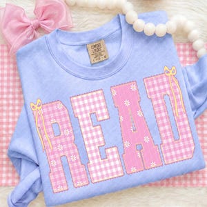 May include: A light blue sweatshirt with the word "READ" in pink and white gingham letters. The letters are decorated with small white flowers and yellow bows. The sweatshirt has a Comfort Colors tag.